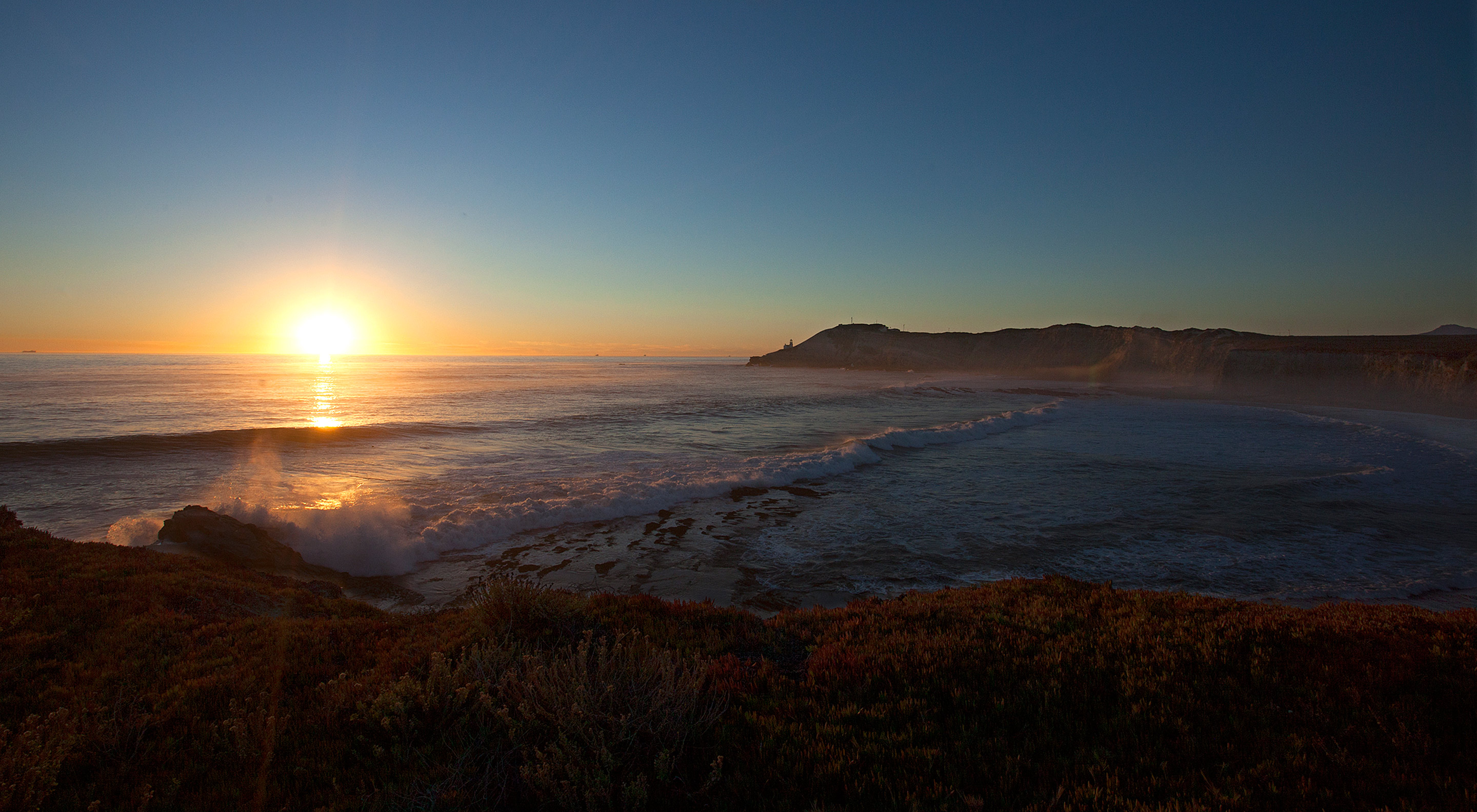 Sun sets over the ocean with gentle waves lapping up against the shore, viewed from a beach.